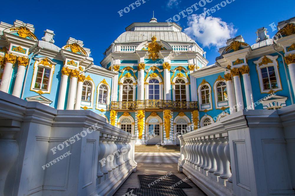 На фотографии изображен центральный фасад Екатерининского дворца в Царском Селе, городе Пушкин, недалеко от Санкт-Петербурга. Богато украшенный золотыми элементами фасад с белыми колоннами гармонично сочетается с ярко-голубым небом. Изящные балконы и декоративные орнаменты подчеркивают стиль русского барокко. Солнечный день делает детали здания особенно выразительными, что привлекает множество туристов со всего мира.